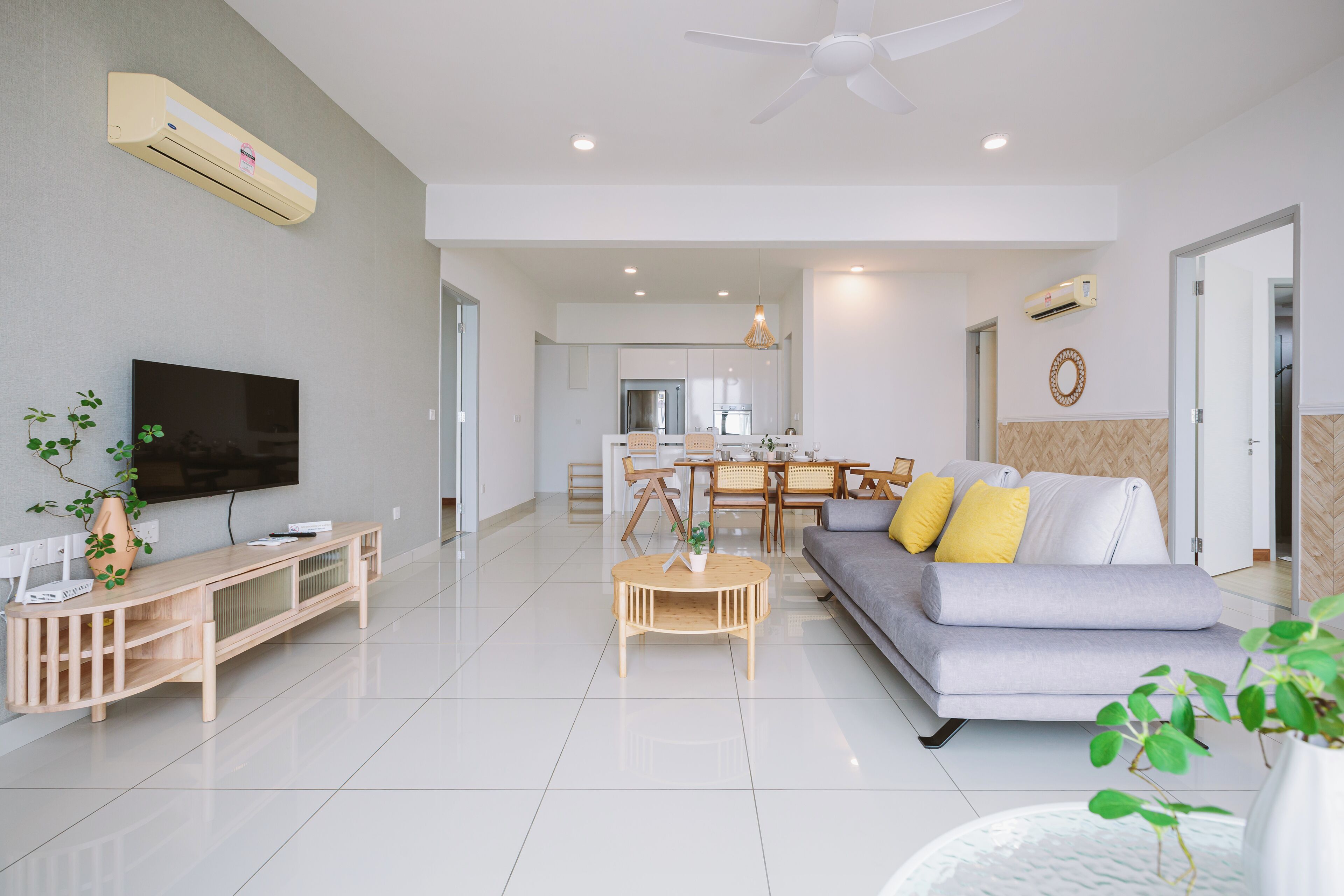 A modern living room with a flat-screen TV, a wooden entertainment unit, a grey sofa with yellow cushions, a round coffee table, and a dining area in the background.