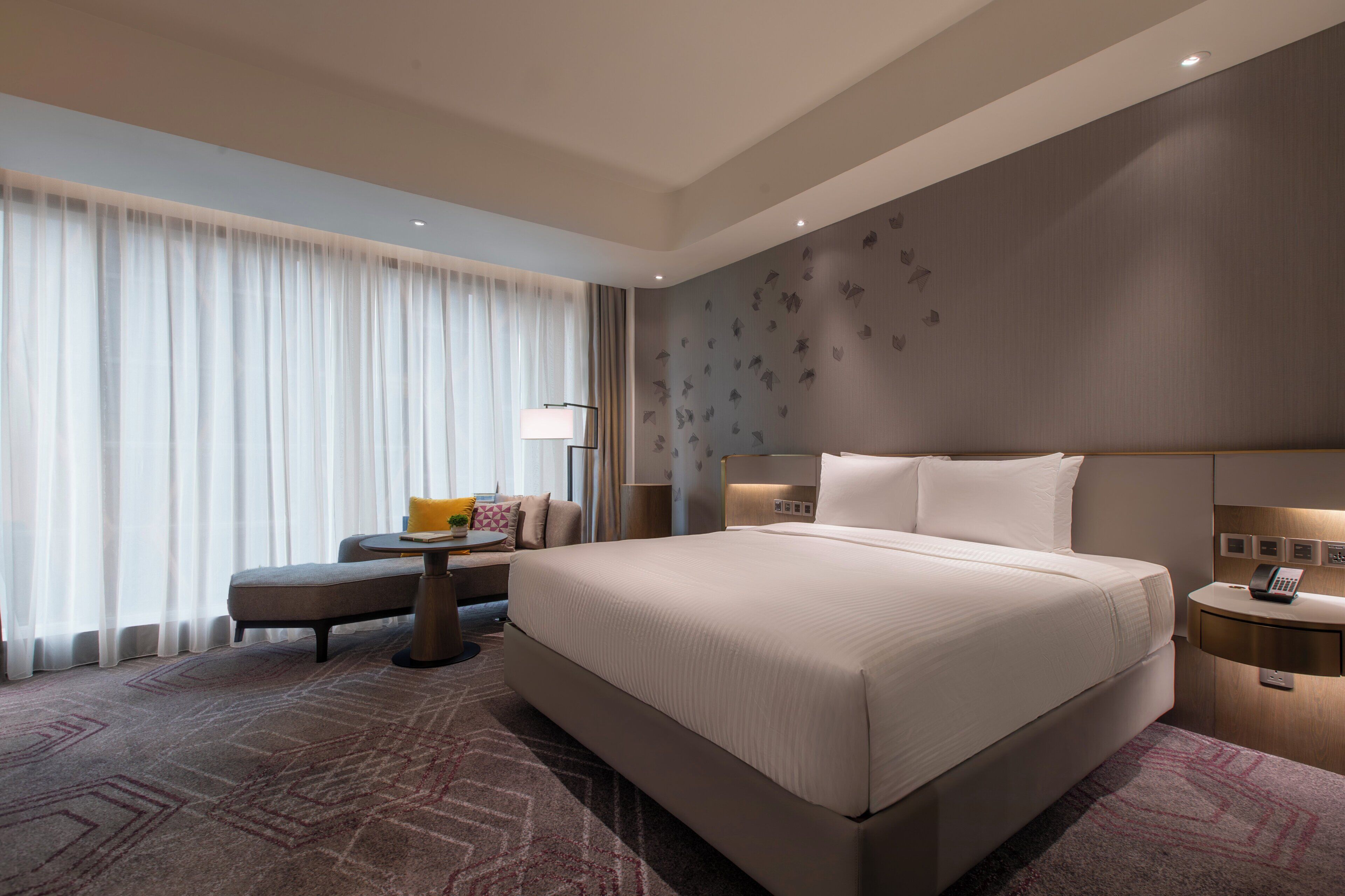 A hotel room with a large bed, a bedside table, a lamp, and a view of the outdoors through sheer curtains.