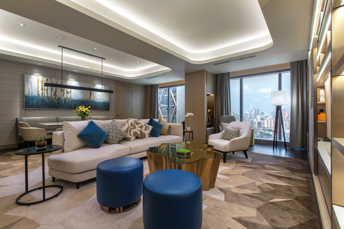 Una sala de estar moderna con un sofá, sillones, una mesa de centro y vistas a la ciudad a través de amplios ventanales.