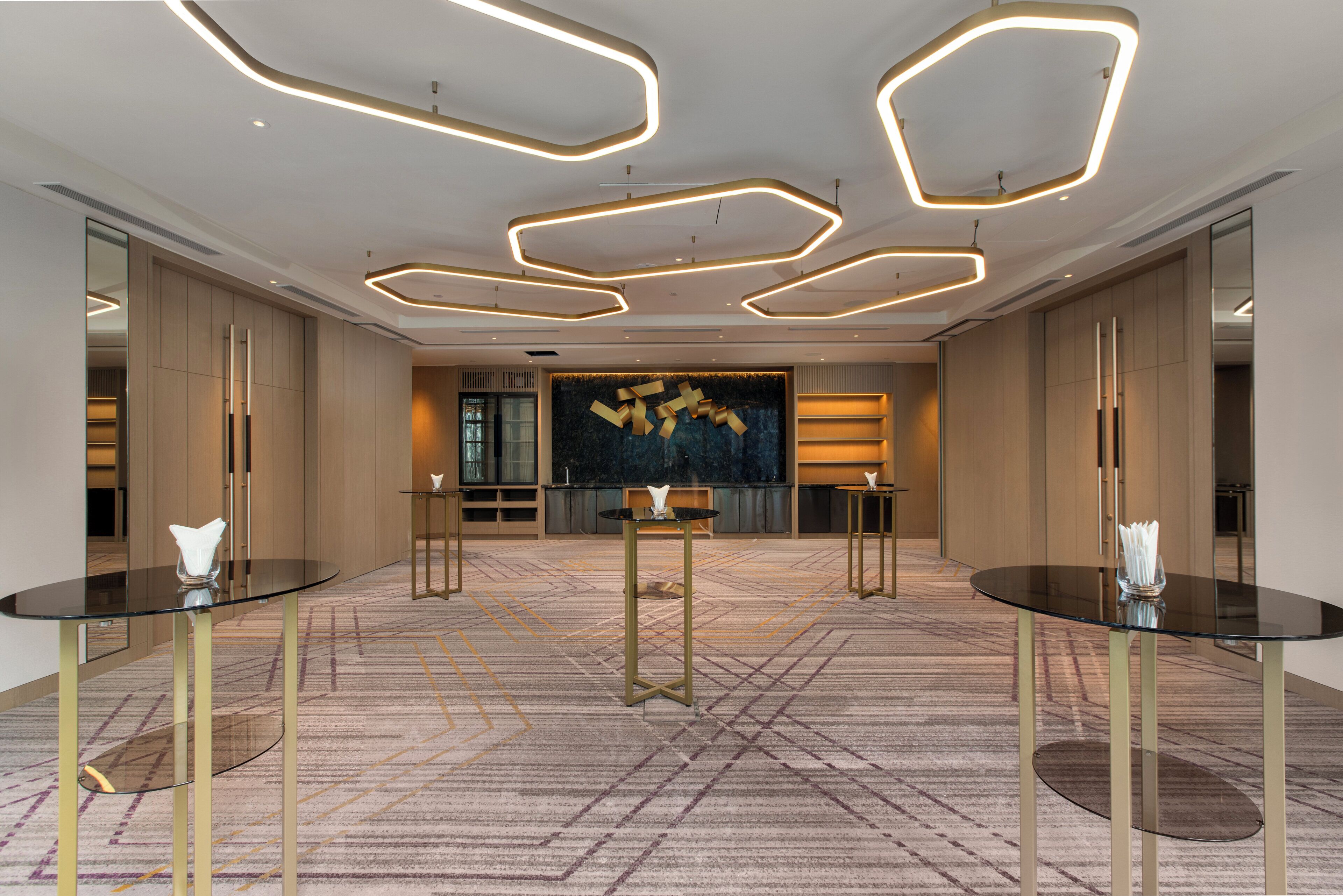 A modern hotel lobby with geometric ceiling lights, a central reception desk, and symmetrical seating arrangements.