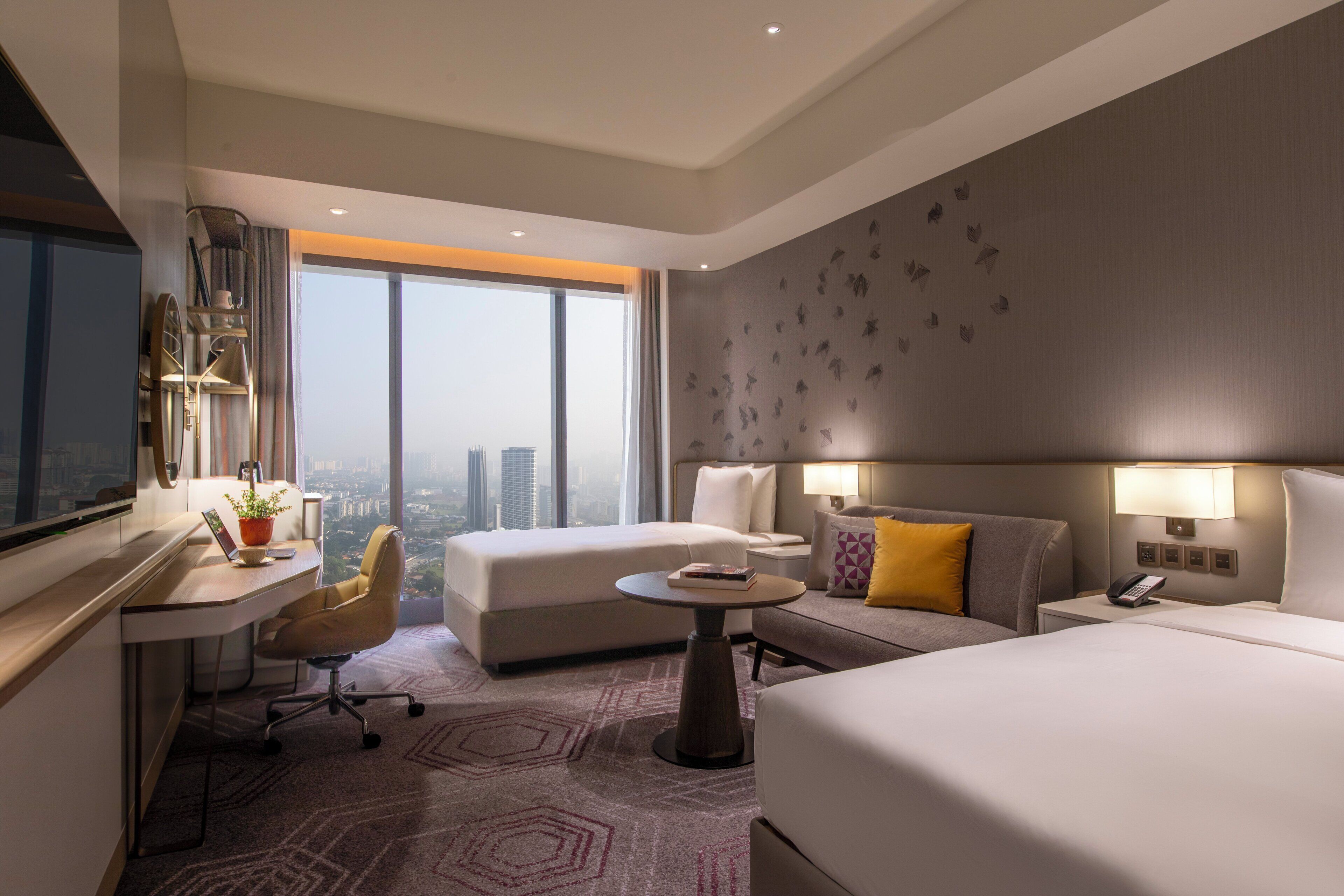 A hotel room with a bed, two sofas, a desk, and a large window offering a city view.