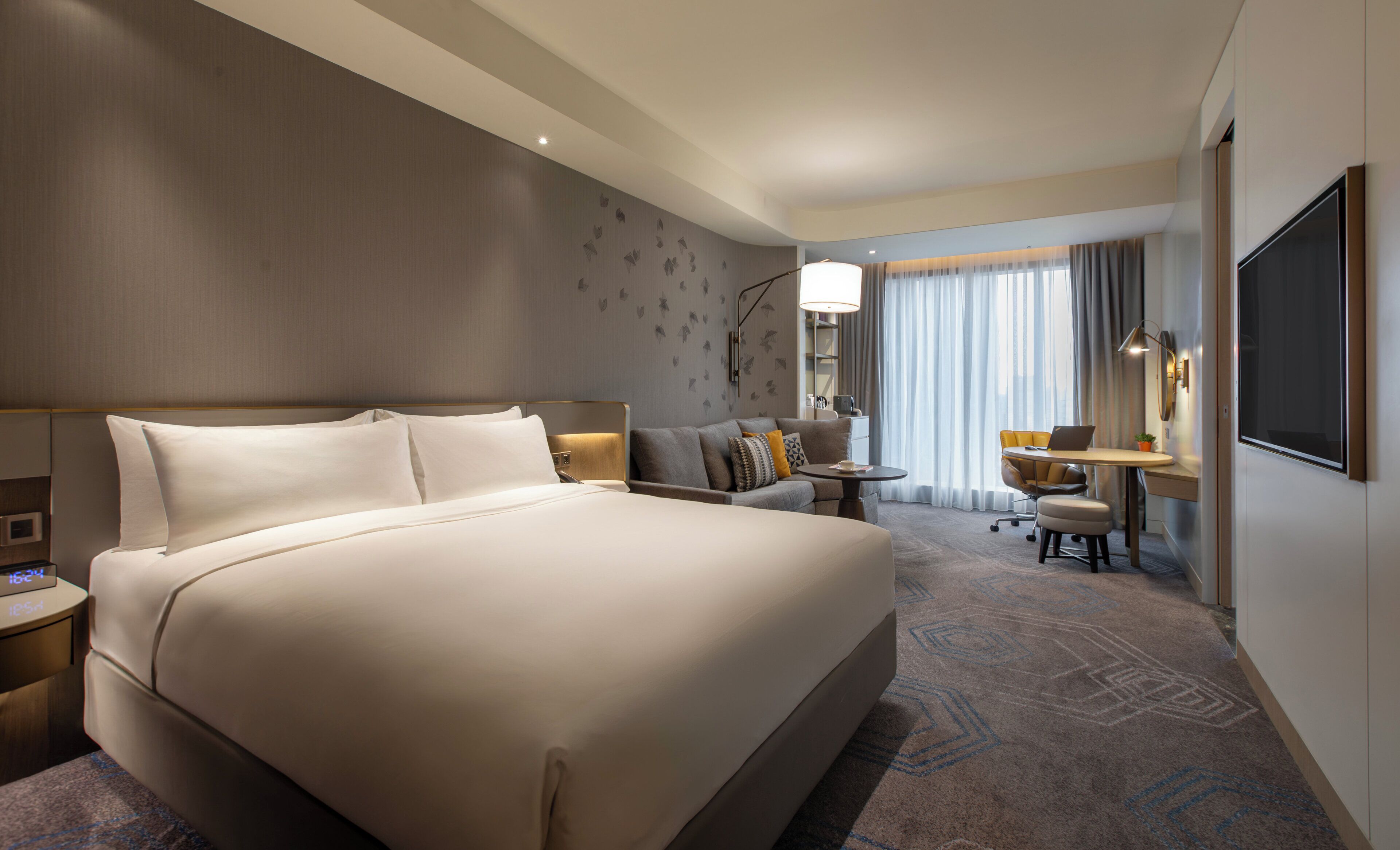 A hotel room with a large bed, a desk, a chair, a lamp, a TV, and a window with curtains.