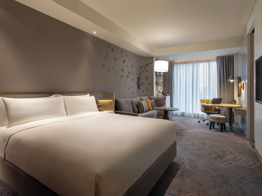 A hotel room with a large bed, a desk, a chair, a lamp, a TV, and a window with curtains.