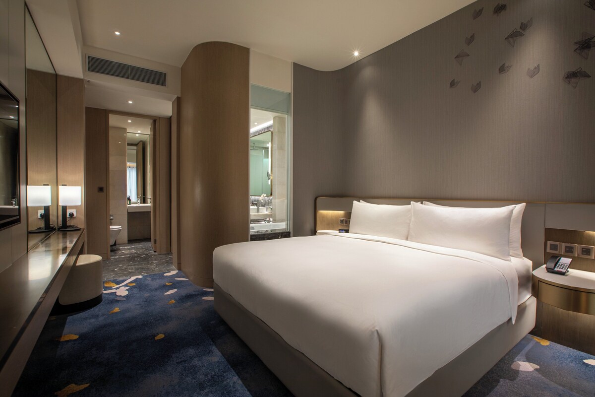Habitación de hotel con una cama grande, una mesita de noche con teléfono, un espejo y un baño visible a través de una puerta abierta.
