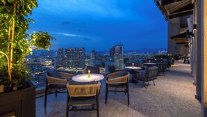 Una terraza al aire libre con mesas y sillas redondas, con vistas a un paisaje urbano al anochecer.
