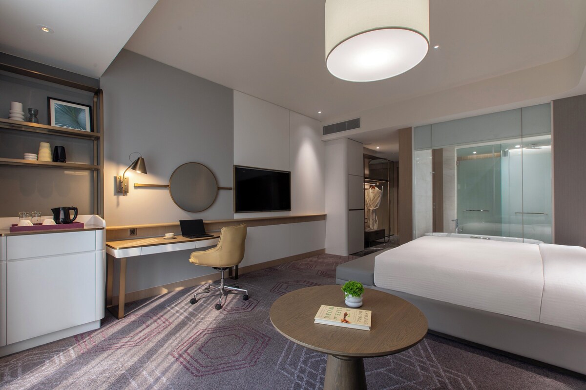 Una habitación de hotel moderna con televisión de pantalla plana, un escritorio con silla, un sofá, una pequeña mesa redonda con una planta y un vestidor.