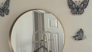 Een ronde spiegel met gouden lijst, een witte deur op de achtergrond en twee vlinderdecoraties aan de muur.