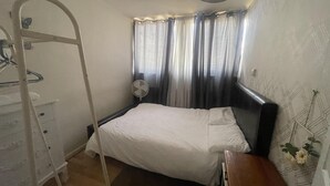 Een slaapkamer met een bed, een nachtkastje, een ventilator, een raam met gordijnen, een radiator en een wandplank met bloemen.