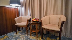 Dos sillones color beige, una mesita redonda con un frutero, una lámpara y un aparador de madera en una habitación con alfombra estampada en azul y amarillo.