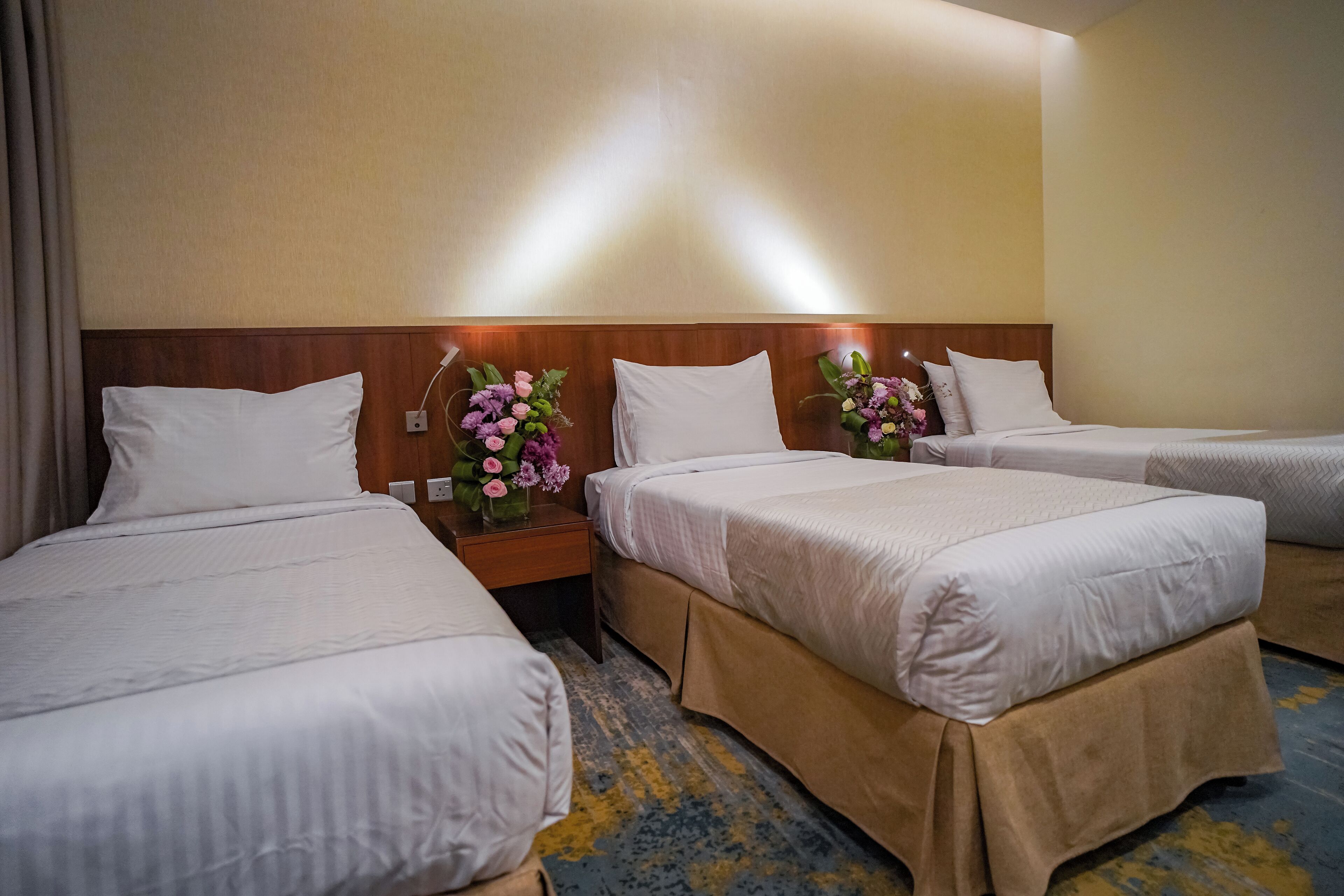 Habitación de hotel con tres camas, cada una con ropa blanca y cabeceros de madera. Hay dos mesitas de noche con centros de flores y una lámpara de pared.