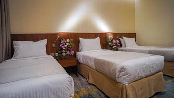 A hotel room with three beds, each with white bedding and a wooden headboard. There are two nightstands with floral centerpieces and a wall-mounted light fixture.