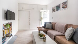 A modern living room with a brown sofa, a white coffee table with decorative items, a bookshelf, and a TV mounted on the wall.