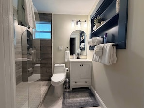 A modern bathroom with a glass shower enclosure, a toilet, a sink with a mirror, and a shelf with towels.