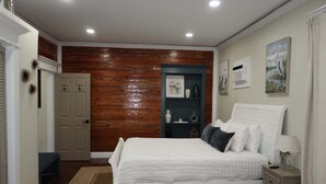 Een slaapkamer met een groot bed, een nachtkastje, een dressoir, een deur, een plank met decoratieve spullen, en een muur met ingelijste foto’s.