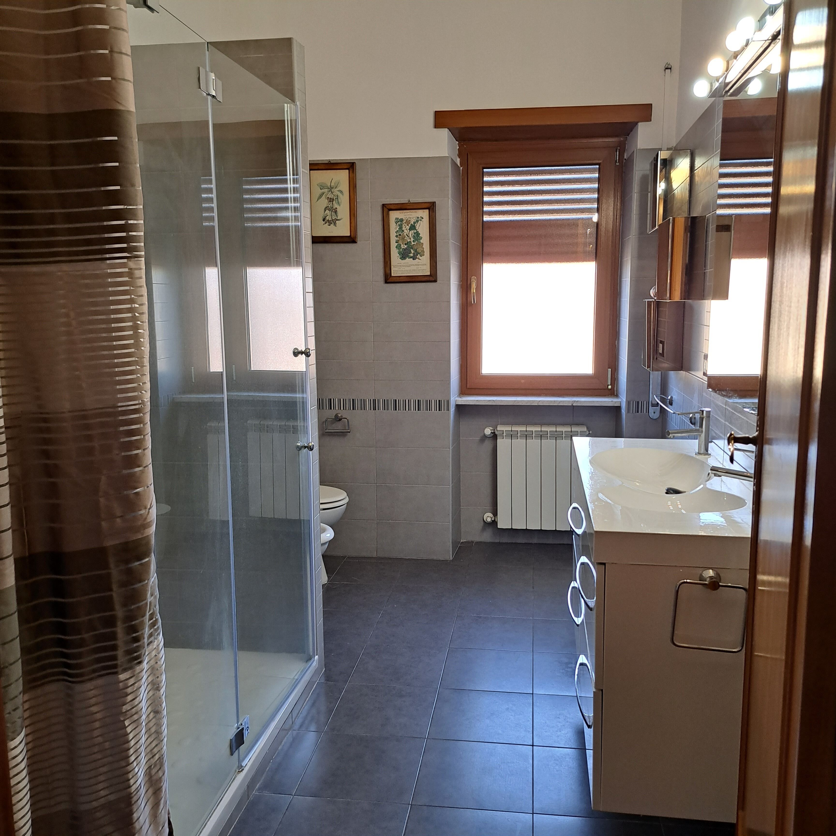 A bathroom with a glass shower enclosure, a toilet, a sink with a mirror, and a window with blinds.