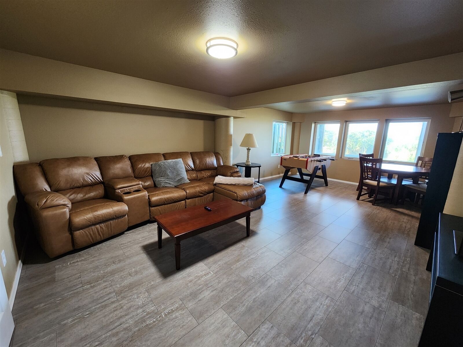 Un salón espacioso con un sofá de cuero marrón, una mesa de centro de madera y una mesa de futbolín. Cuenta con una zona de comedor con mesa y sillas, y grandes ventanales que dejan entrar luz natural.