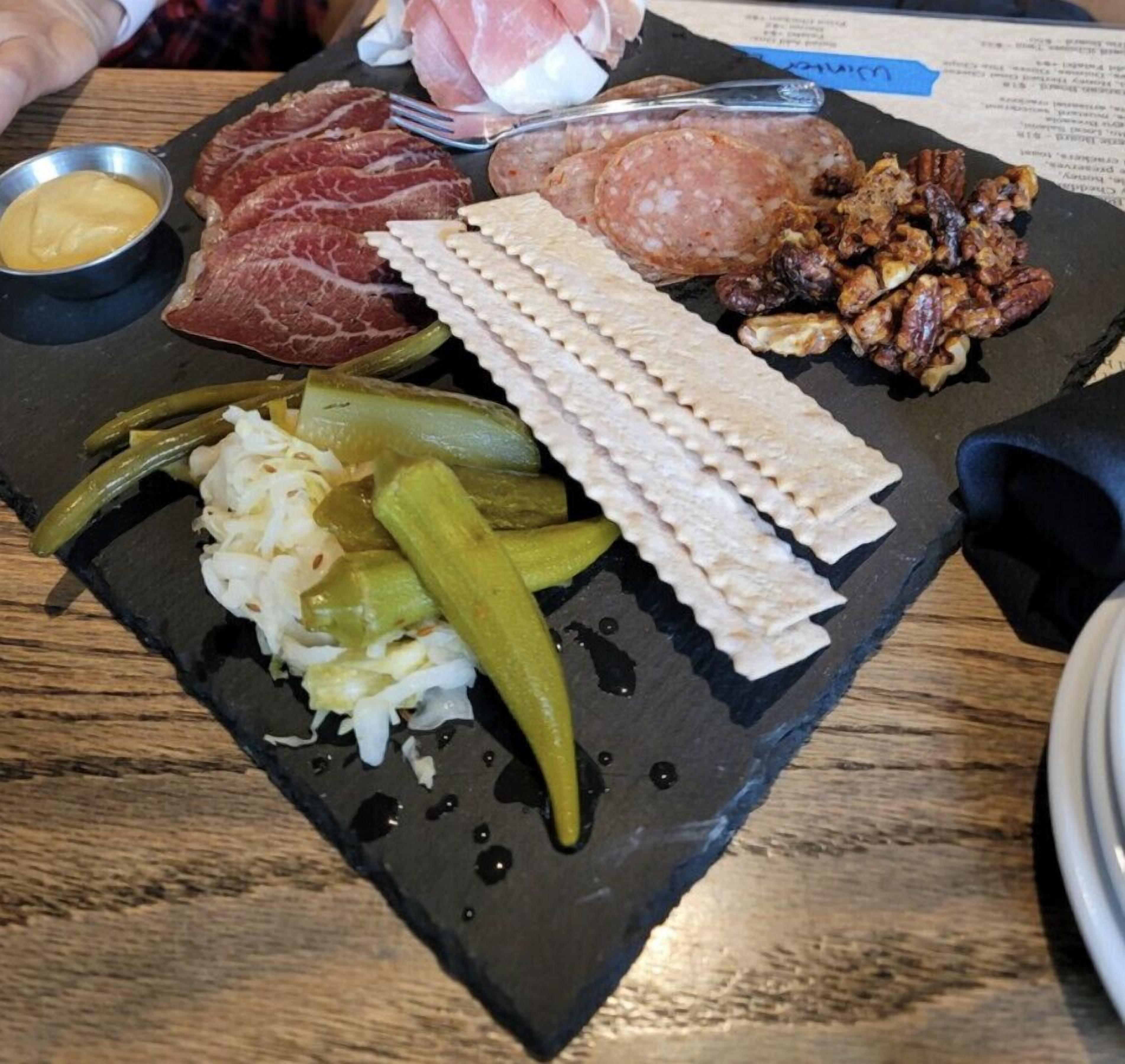 Una tabla de pizarra negra con diferentes carnes, pepinillos y un recipiente pequeño con mostaza.