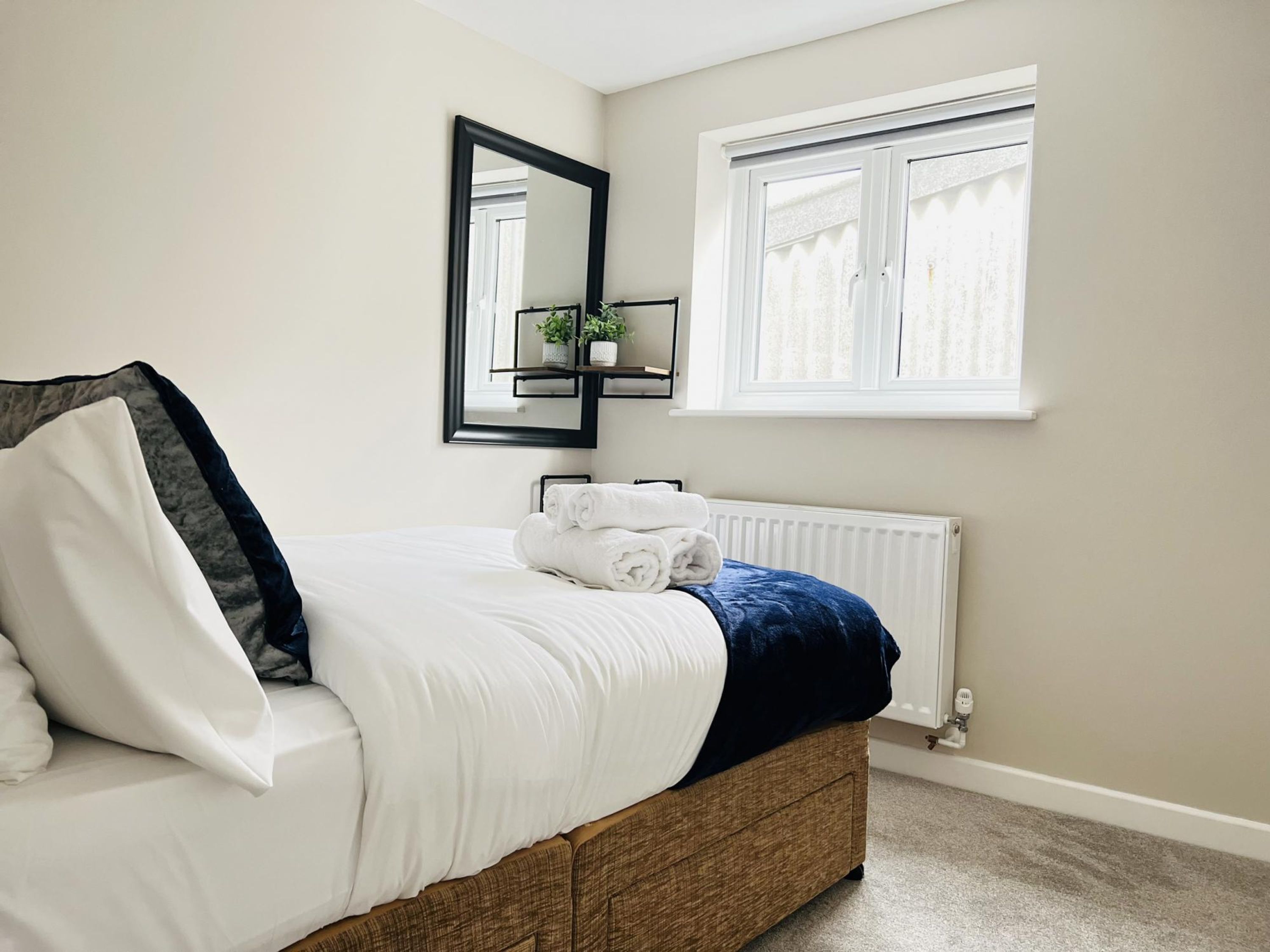 A neatly made bed with white and blue bedding, a mirror above it, and a window with blinds.