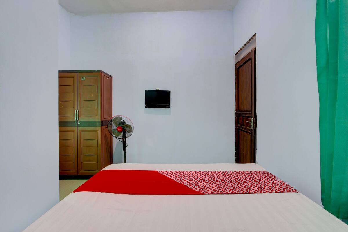 A bedroom with a bed, a wooden wardrobe, a wall-mounted TV, and a fan.