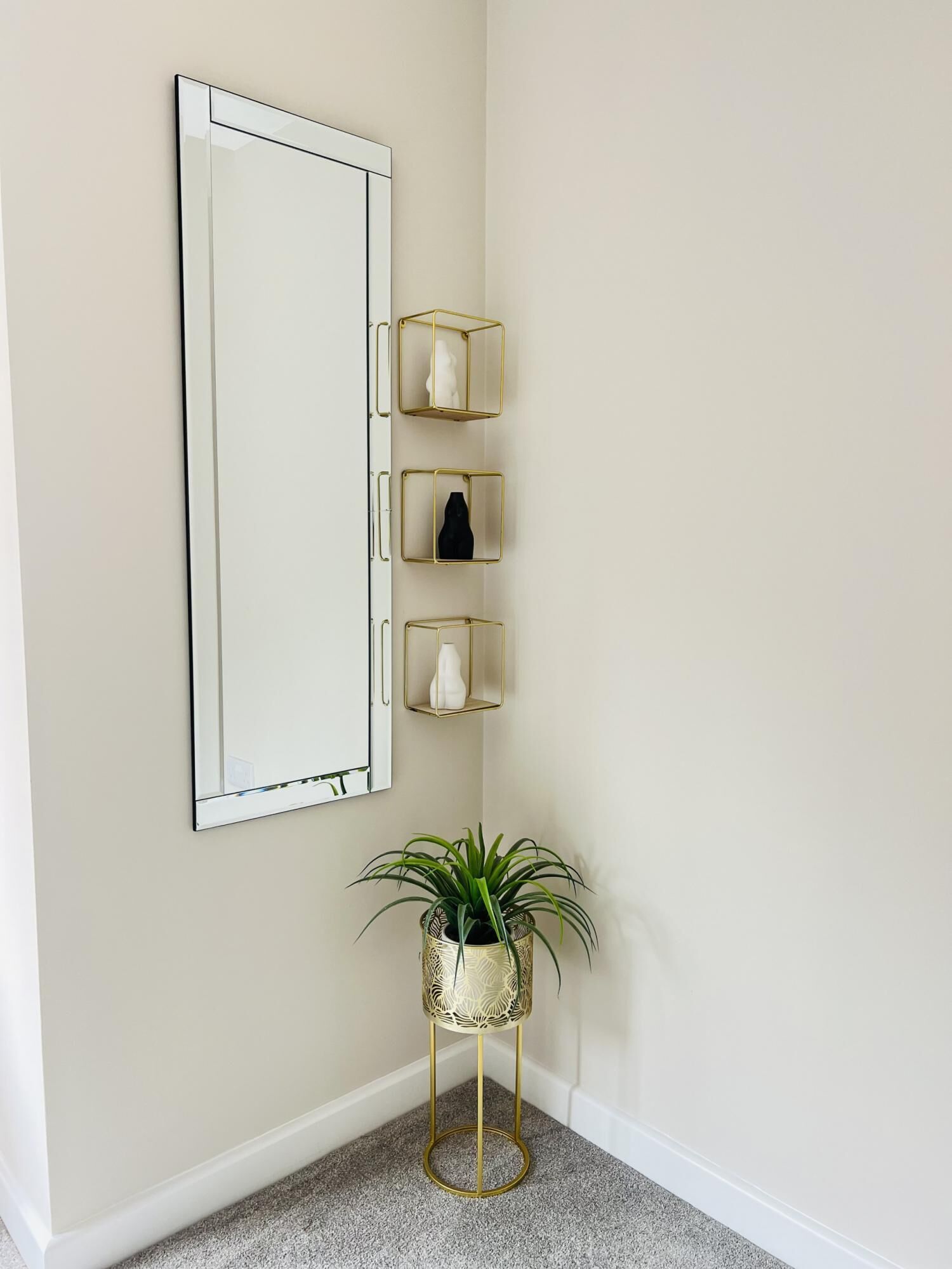 Un rincón con un espejo grande, tres estantes dorados con objetos decorativos y una planta en maceta sobre un pedestal dorado.