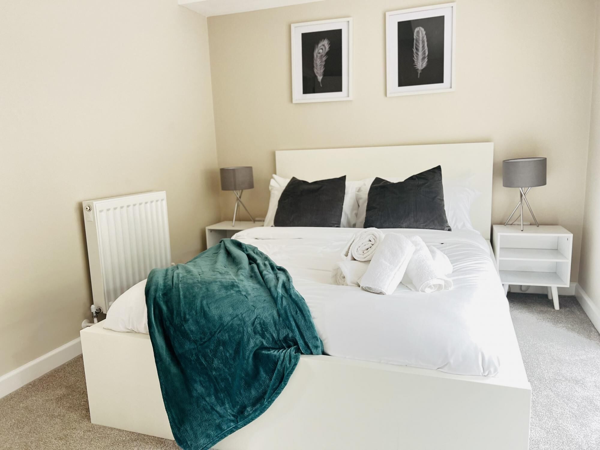 A neatly made bed with white linens and a teal throw blanket, two bedside lamps, a white nightstand, and two framed feather art pieces on the wall.