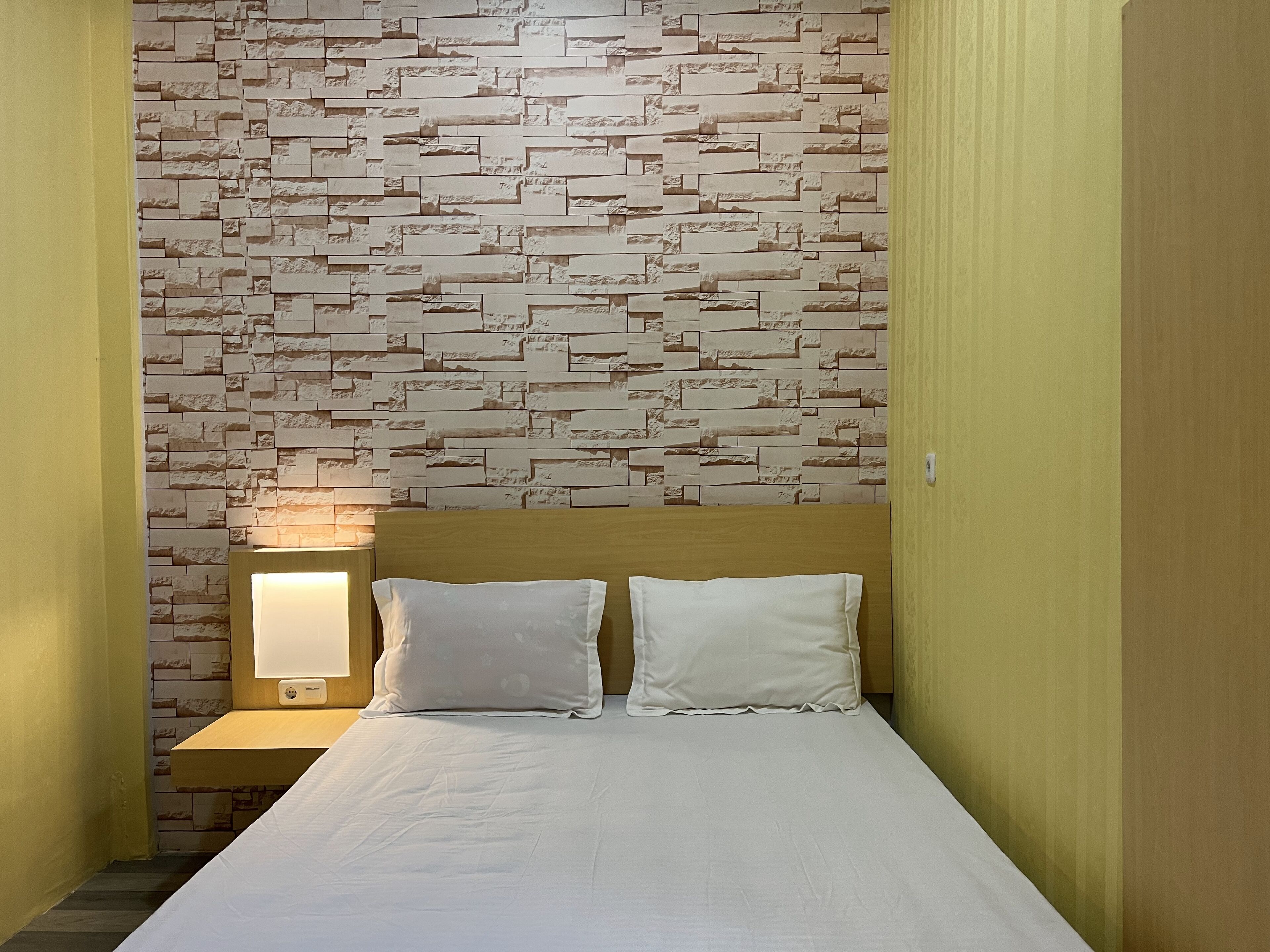 A hotel room with a bed, two pillows, a wooden headboard, a nightstand with a lamp, and a textured wall.