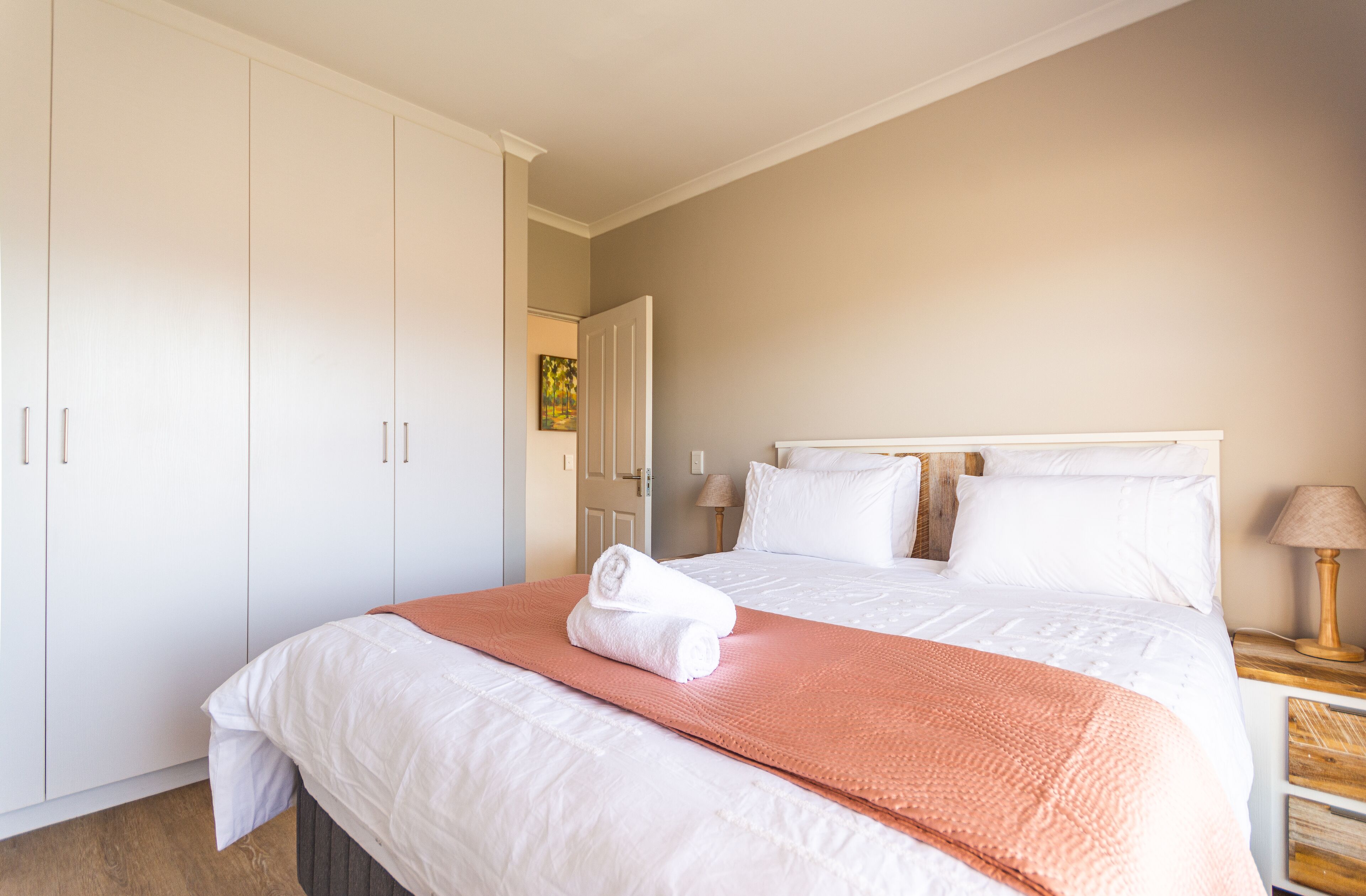A bedroom with a large bed, white bedding, a pink throw blanket, and a wooden nightstand with a lamp.