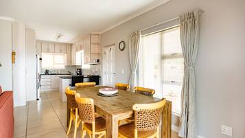 A spacious dining area with a wooden table and wicker chairs, a kitchen in the background, and a large window with curtains.