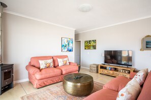 A living room with a pink sofa, a round coffee table, a TV on a wooden stand, and a fireplace. - Capri House (Cape Town)