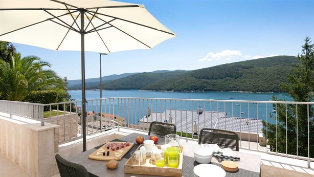 Ein Balkon mit gedecktem Tisch für das Frühstück, mit Blick auf einen See, auf dem Boote fahren, und Hügel im Hintergrund.