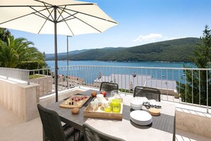 Un balcón con una mesa dispuesta para el desayuno, con vista a un lago con barcos y colinas al fondo.