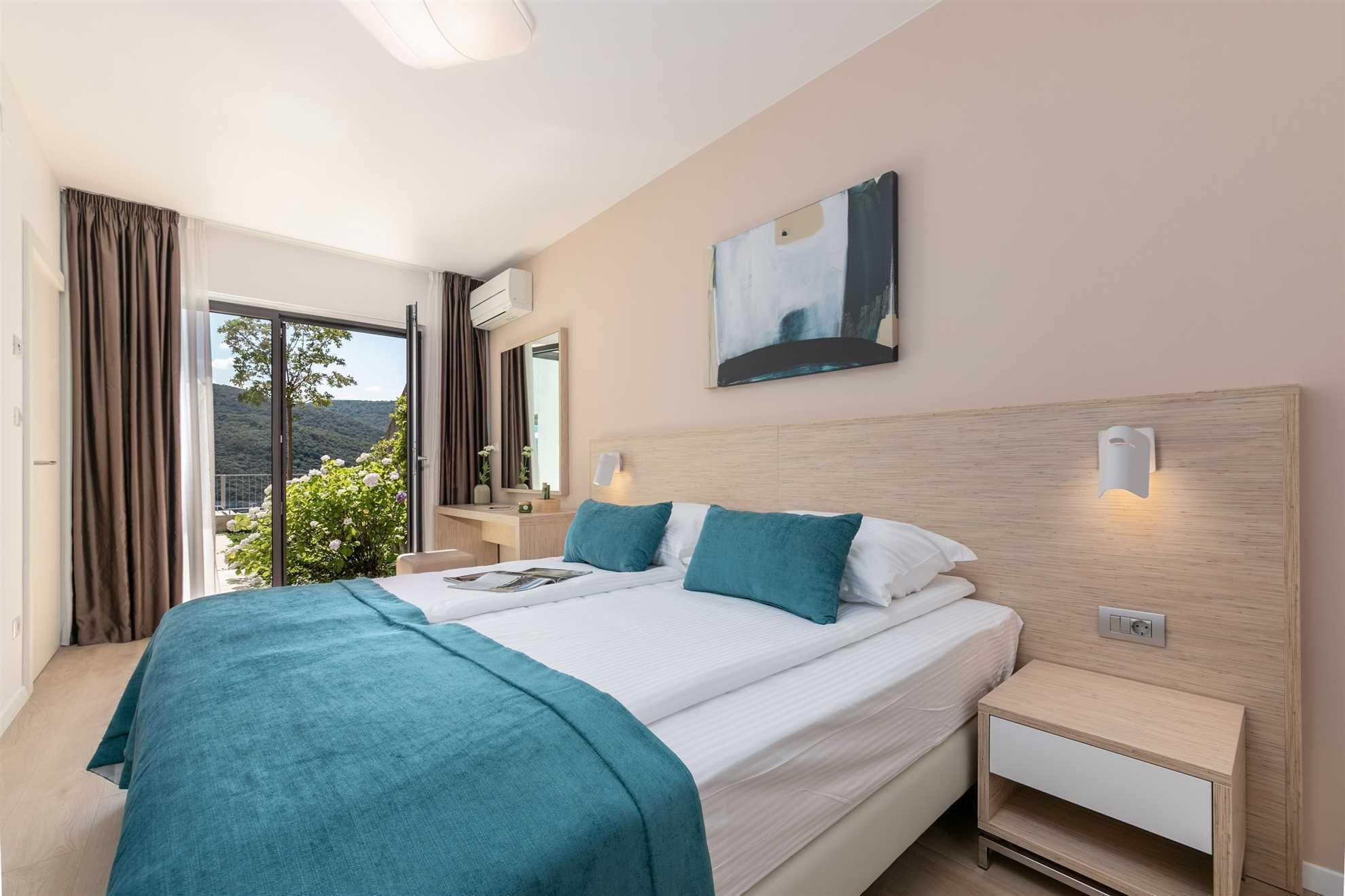 A modern hotel room with a large bed, bedside tables, a nightstand, a painting, and a view of greenery.