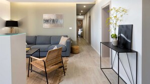 Un salon moderne avec un canapé gris, un fauteuil en osier, une table basse en verre et un meuble TV noir agrémenté d’une plante et d’un tableau.