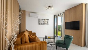 Een moderne woonkamer met een bank, een groene fauteuil, een flatscreen-televisie en een groot raam met uitzicht op de omgeving.