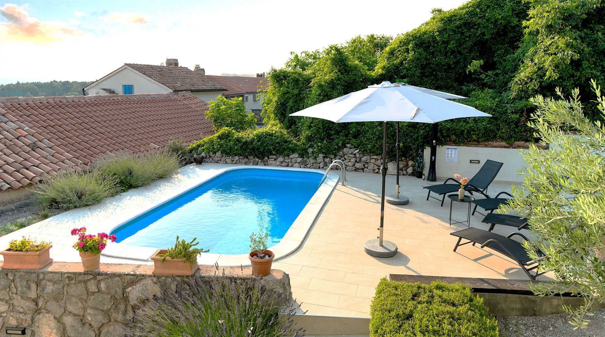 A swimming pool with a tiled deck, surrounded by potted plants and a lounge area with chairs and an umbrella.