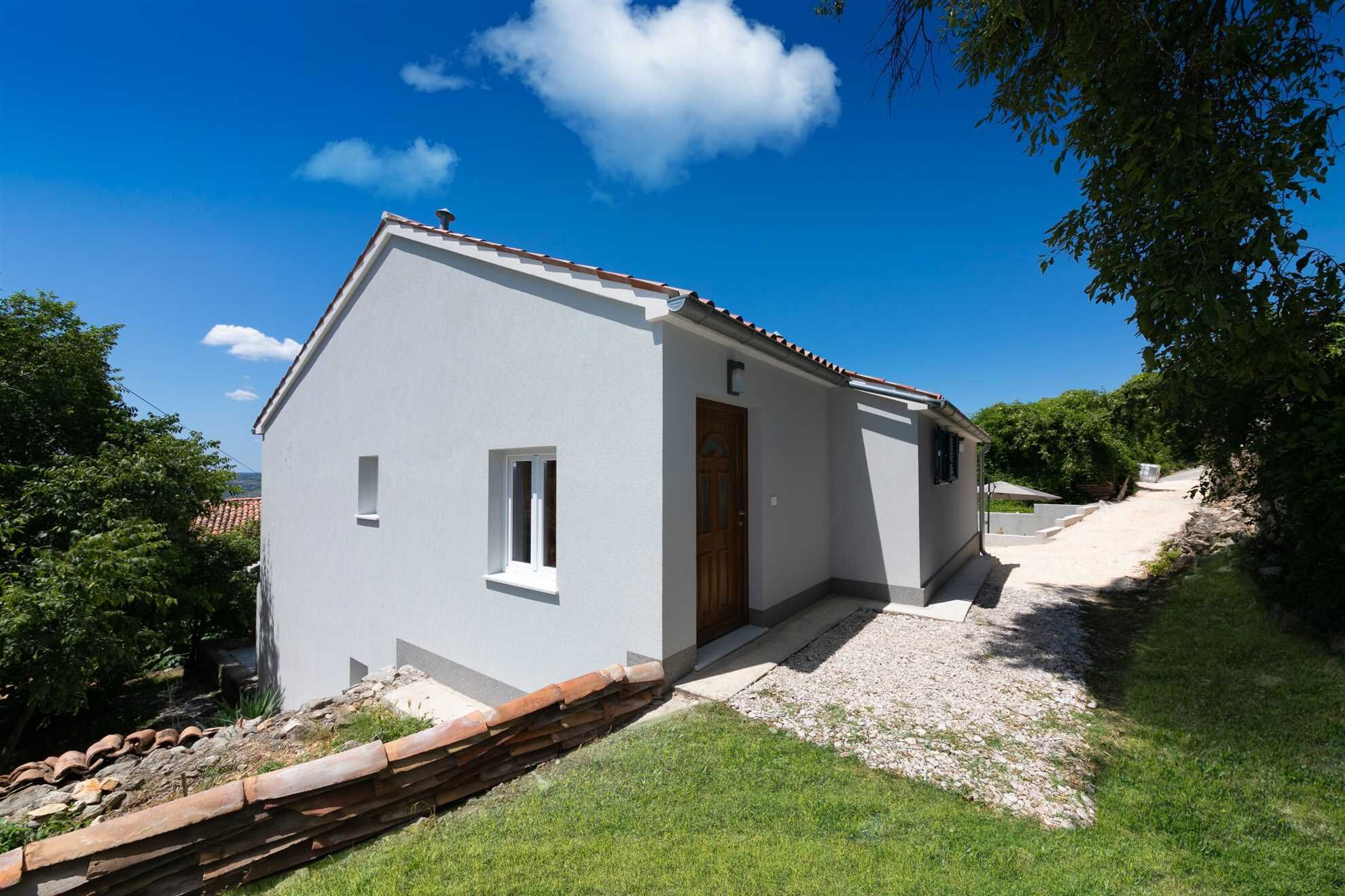 Um edifício térreo branco com porta marrom e uma pequena janela, rodeado por vegetação e um caminho de cascalho.