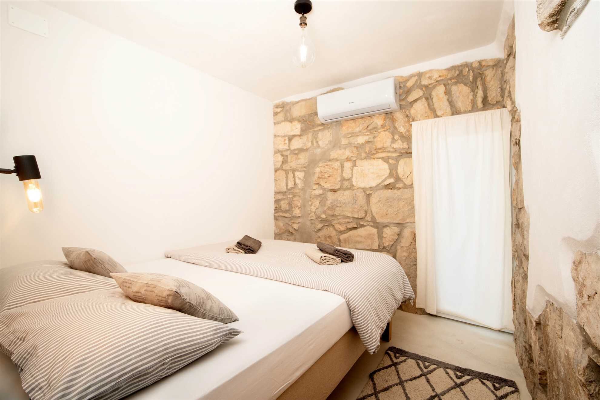 A bedroom with two beds, a stone wall, an air conditioning unit, and a window with curtains.