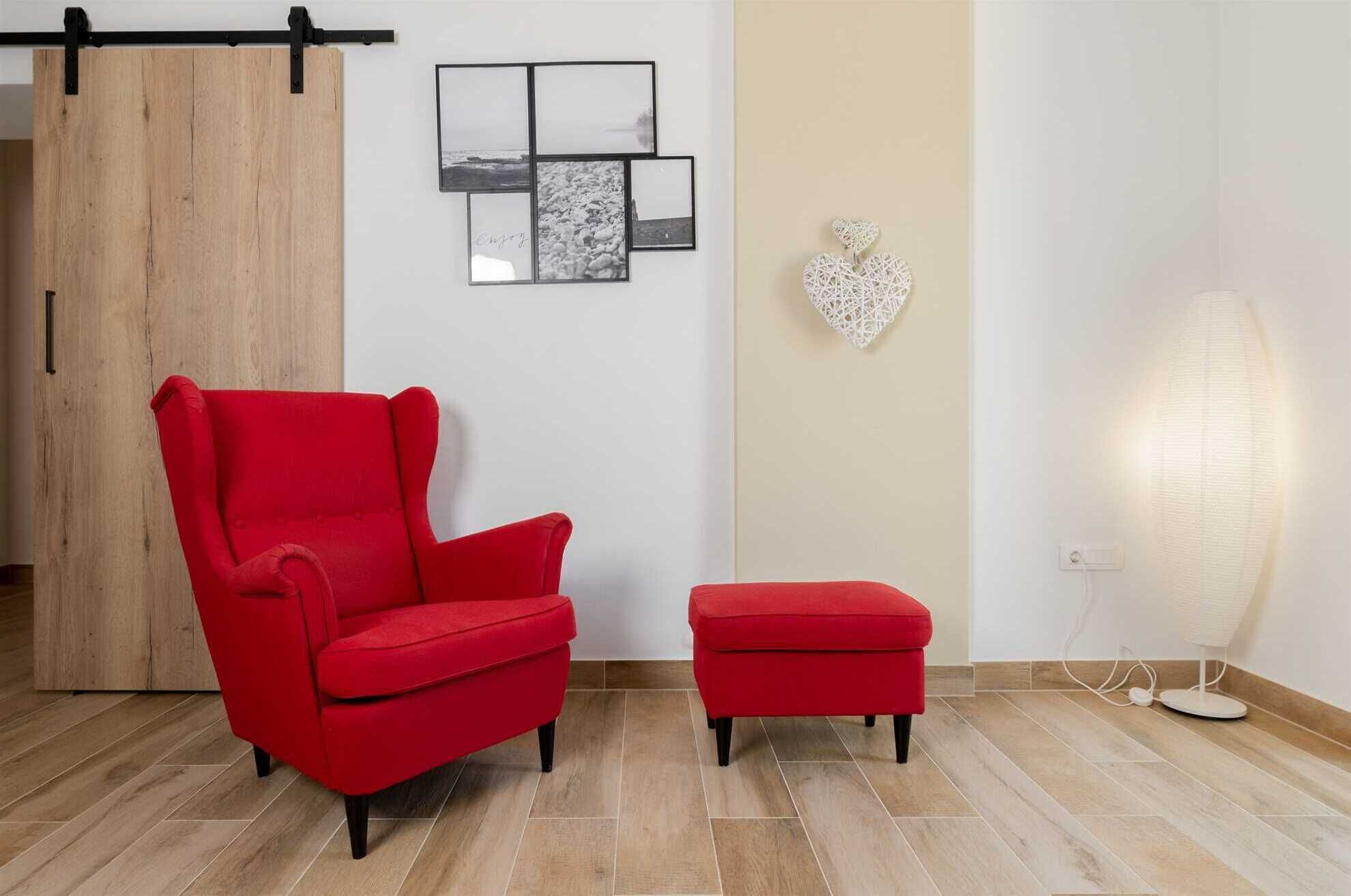 A room with a red armchair and ottoman, a wooden sliding door, and a wall with framed pictures and a heart-shaped decoration.
