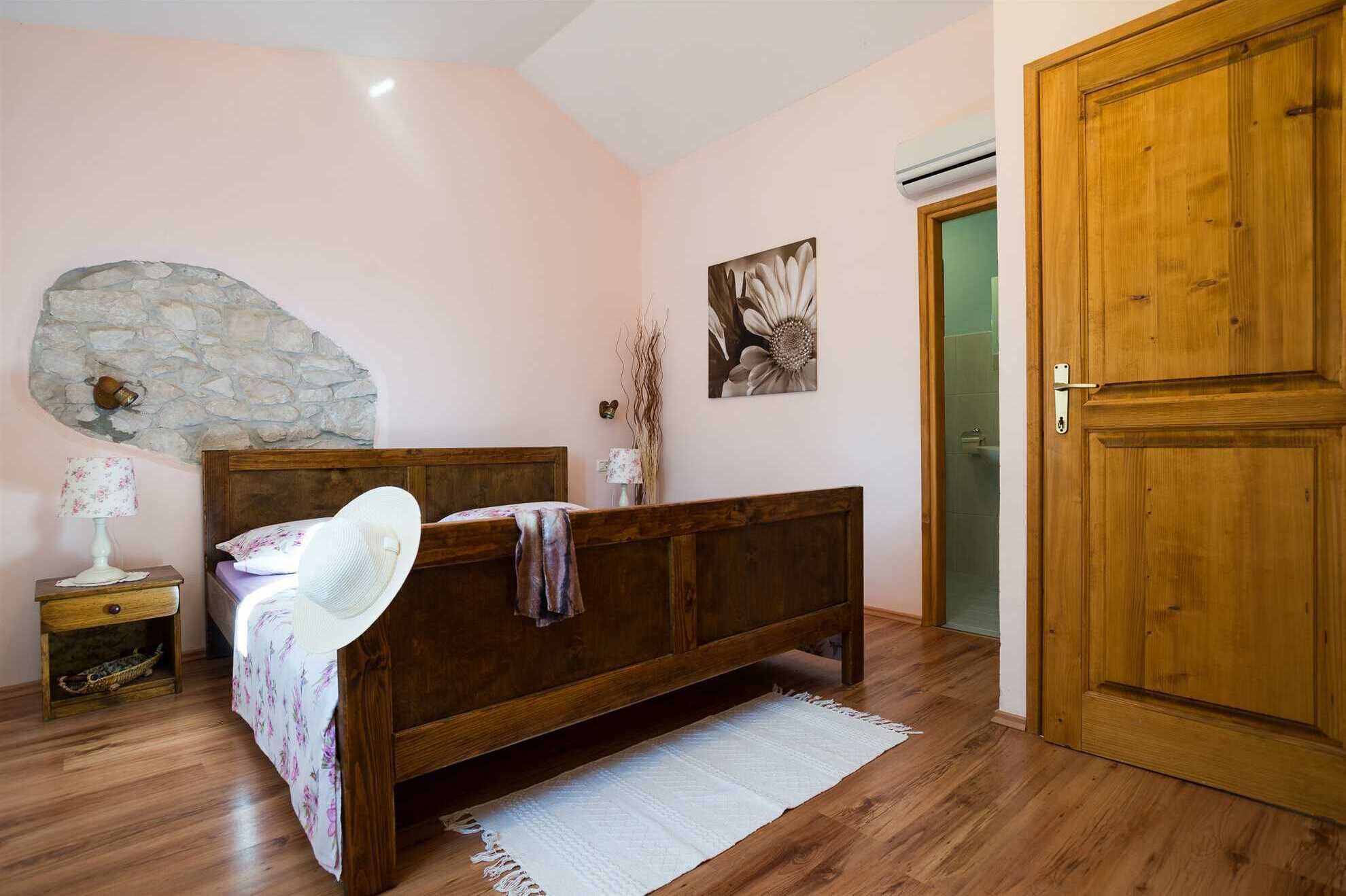 A bedroom with a wooden bed, a stone wall, a wooden door, and a small bedside table with a lamp.