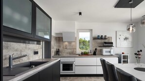 A modern kitchen with dark cabinetry, a large window, and a dining area with a table and chairs. - Mediterranean villa Anatai with a sea view (Drenje)