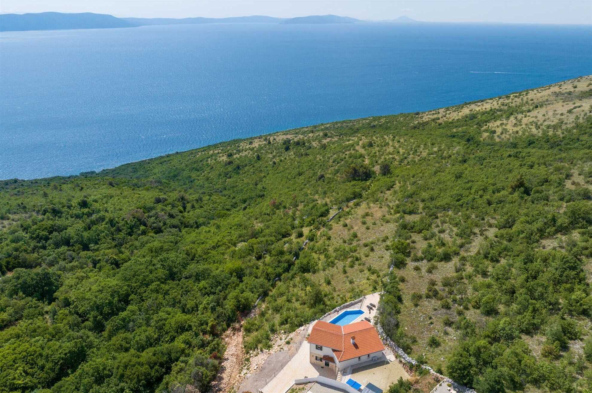 An aerial view of a coastal property with a swimming pool, surrounded by dense greenery and overlooking the sea.