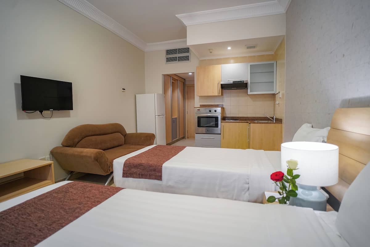 Hotellihuone, jossa on kaksi sänkyä, televisio, sohva, pieni keittiö ja ruusu yöpöydällä.