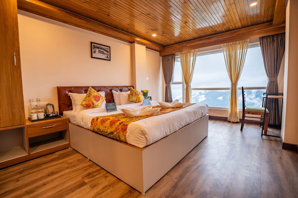 A bedroom with a large bed, wooden headboard, a nightstand with a coffee maker, and a view of the sky through the window.