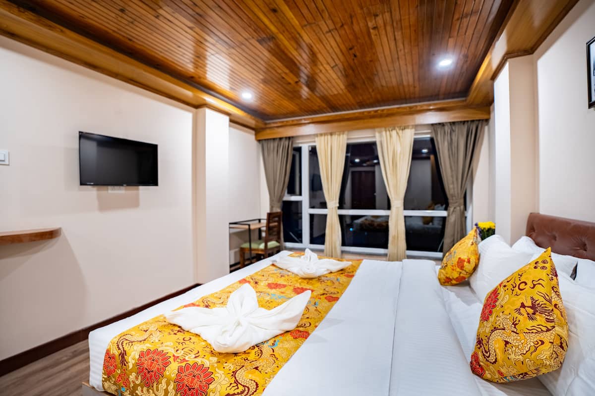 A hotel room with a wooden ceiling, a flat-screen TV mounted on the wall, a bed with a yellow patterned bedspread and white pillows, and a window with curtains.