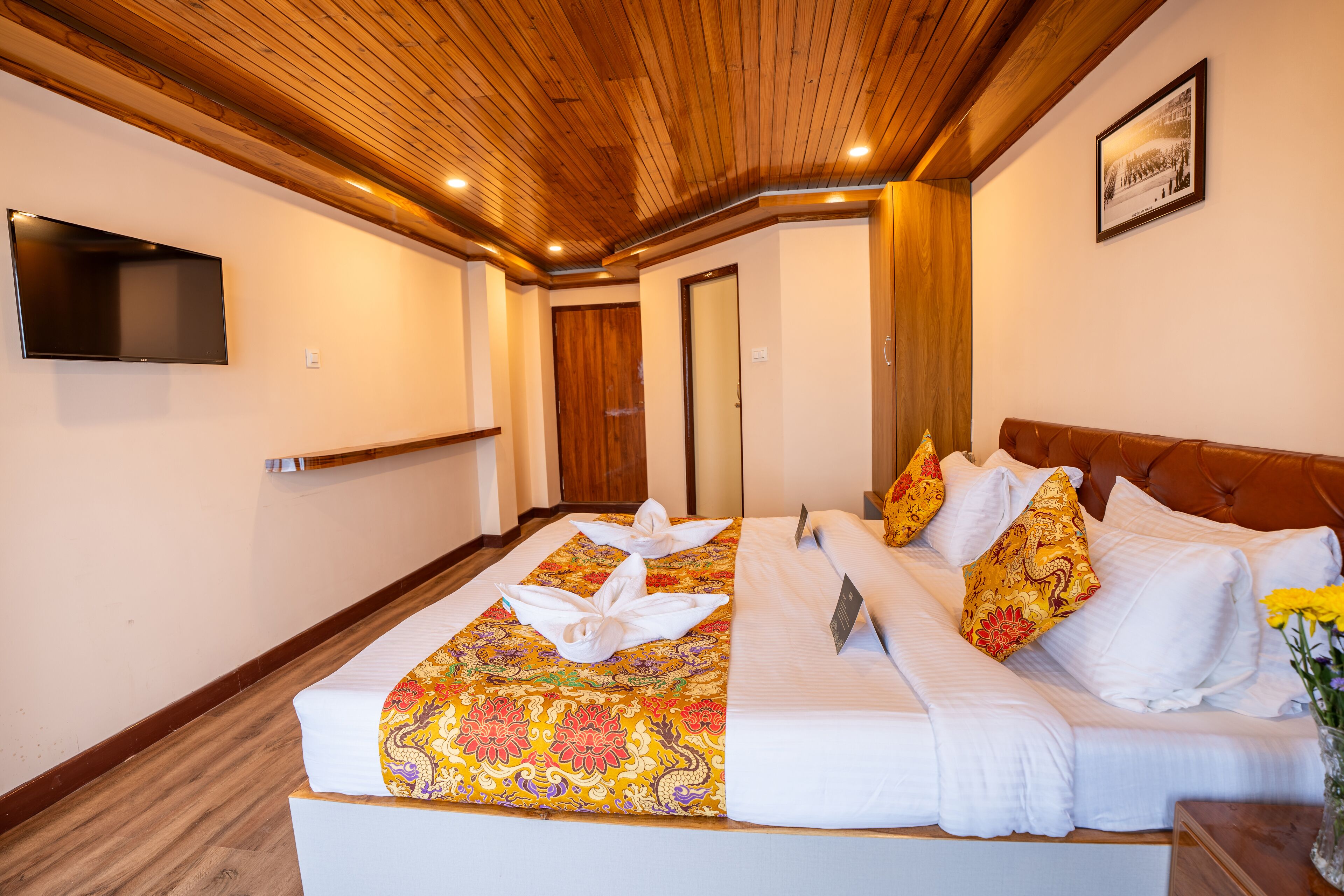 A hotel room with a wooden ceiling, a bed with a floral bedspread, white pillows, and a flat-screen TV mounted on the wall.