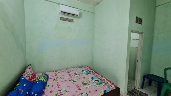 A small, single-bed room with a pink bedspread featuring cartoon characters, a green chair, and a white air conditioning unit mounted on the wall. - SPOT ON 93020 Rumah Singgah Ganjuran (Sumbermulyo)