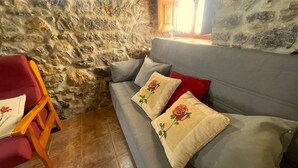 A stone wall, a grey sofa with rose-patterned pillows, a wooden chair, and a red pillow. - Casa El Collau (Sevares)