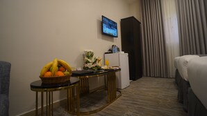 A hotel room with a bed, a television mounted on the wall, a small table with a fruit basket, a vase with flowers, and a refrigerator. - Emaar Andalusia (Makkah)