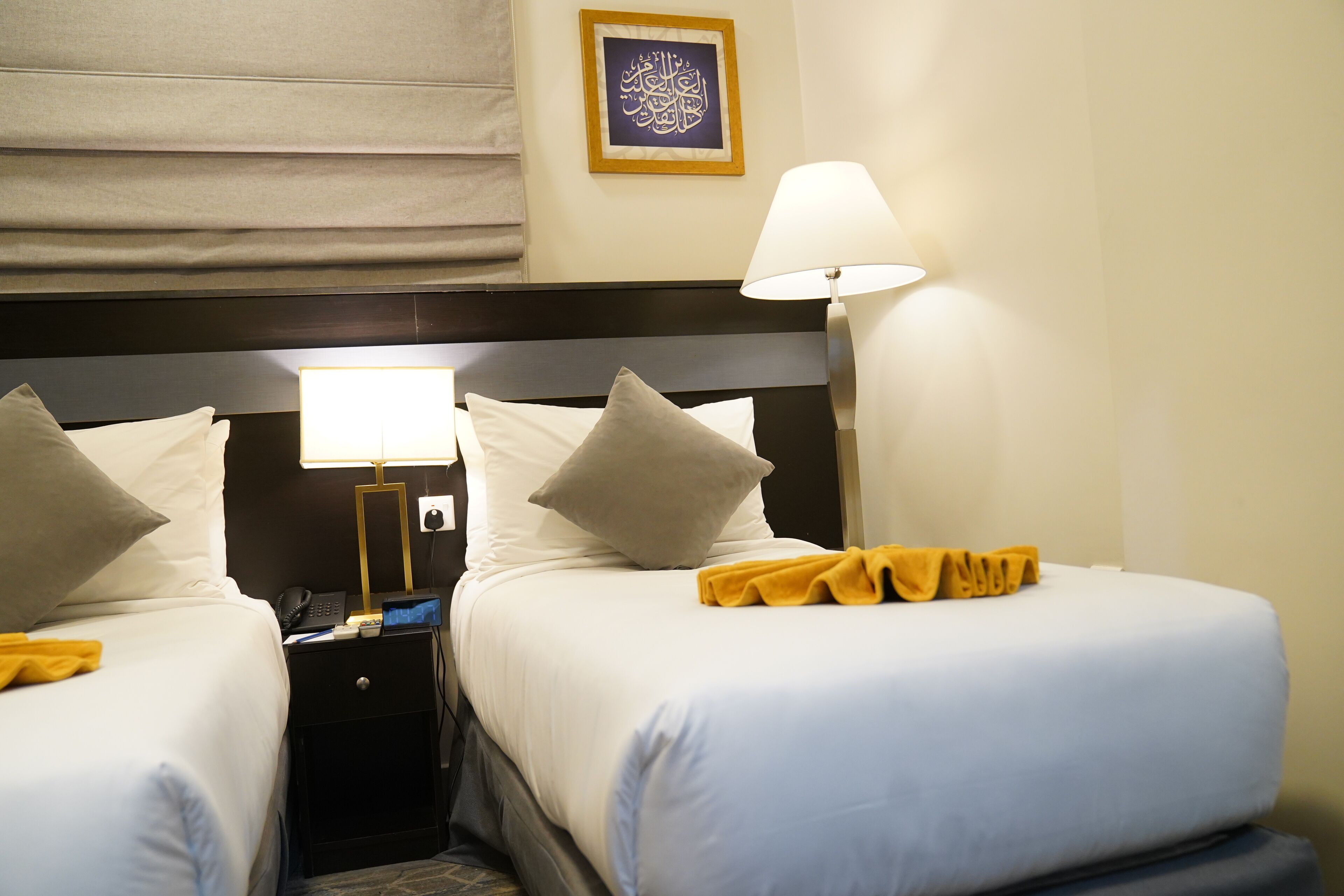 A hotel room with two beds, each with a pillow, a bedside table with a lamp, and a small nightstand with a phone and a laptop.