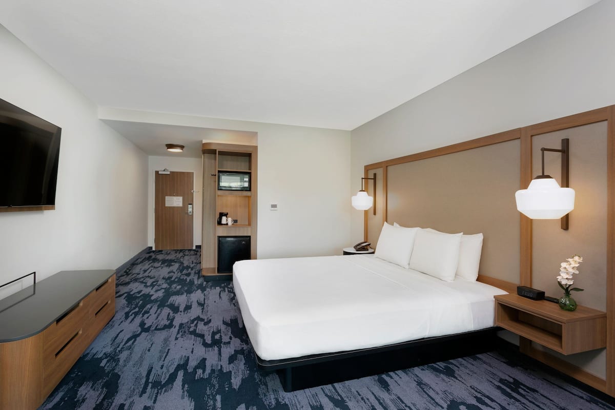 A hotel room with a large bed, a flat-screen TV mounted on the wall, a desk with drawers, a small kitchenette, and a bedside table with a lamp and a vase.